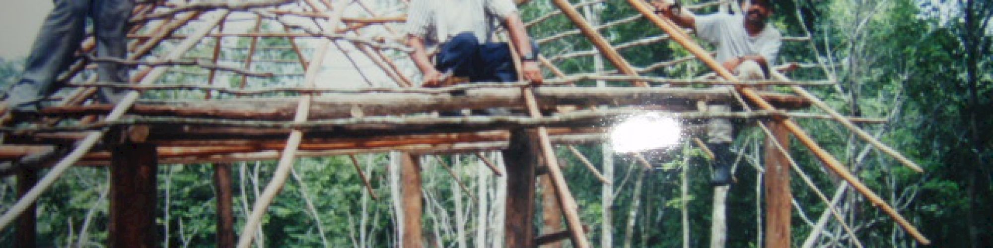 Three men are constructing a wooden structure in a forested area, using logs and branches for the framework.