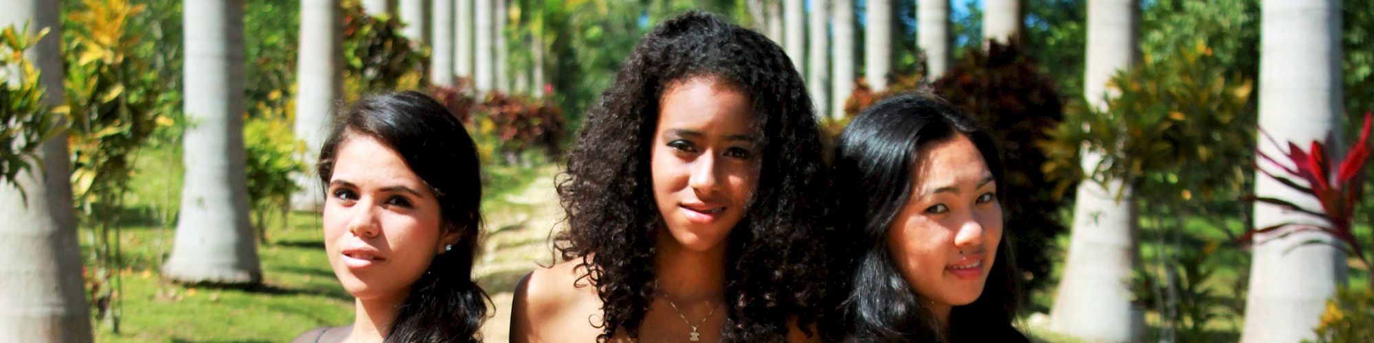 This image shows three young women standing outdoors in a lush area with palm trees lining the background, posing and looking at the camera.