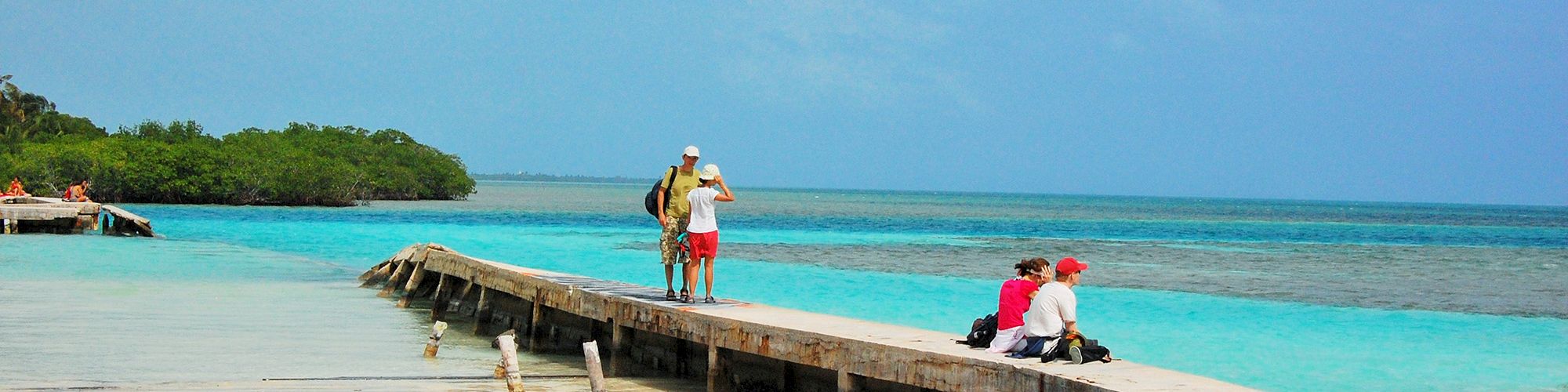 A sandy beach with a pier extending into turquoise waters; people are sitting and walking on the pier under a partly cloudy sky.