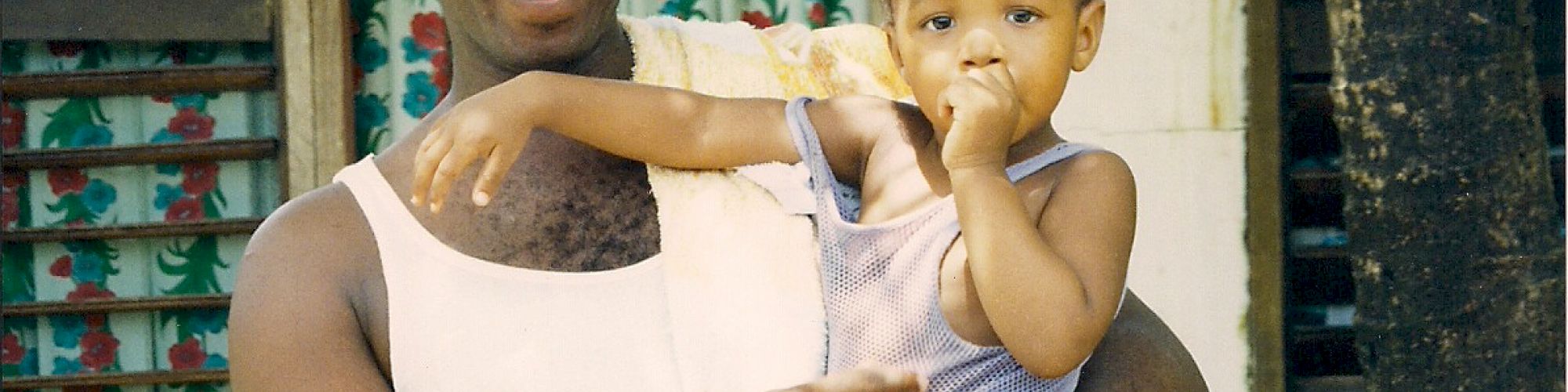 A man in a white tank top holds a child outside a house with colorful curtains, smiling under the shade of a tree.