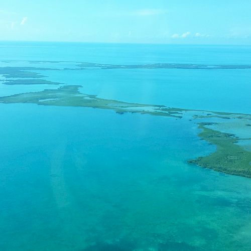 This image shows a series of small, elongated islands and clear blue waters under a vibrant blue sky. The islands are surrounded by shallow, turquoise water.