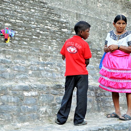 A boy in a red shirt and a woman in traditional attire are standing on stone steps, engaged in conversation.