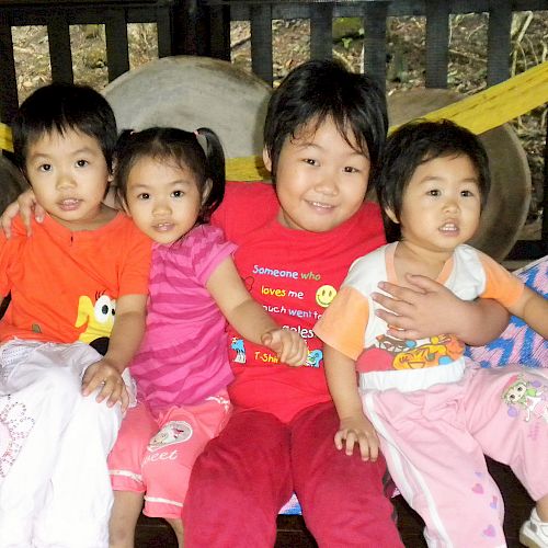 Four young children are sitting close together on a hammock, smiling at the camera, with a colorful background behind them.