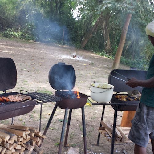 A person is grilling food outdoors using three barbecues, with smoke rising from the grills. A pot and pile of firewood are nearby.