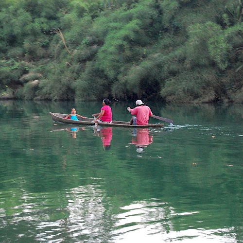 A small boat with three people is on a calm river surrounded by lush green trees, reflecting in the water, while they paddle.