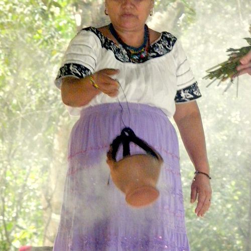 A woman wearing traditional clothing performs a ritual with a smoking pot, in an outdoor setting surrounded by greenery.