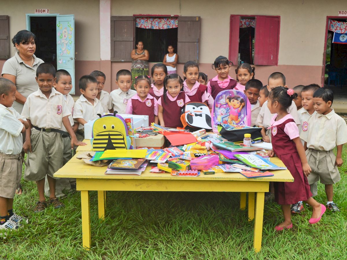 A group of children in school uniforms stand around a table filled with school supplies and backpacks, outdoors, with a teacher nearby.