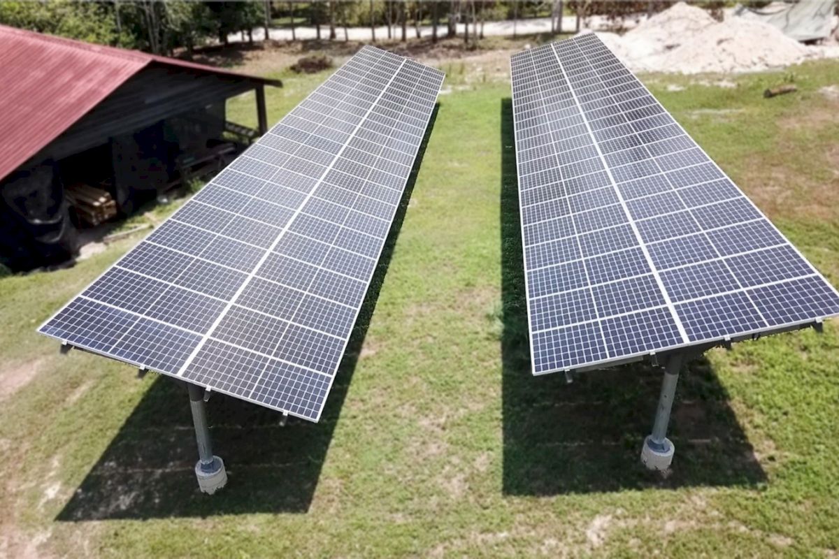 The image shows two large solar panel arrays installed outdoors on a grassy area, with a small building featuring a red roof in the background.