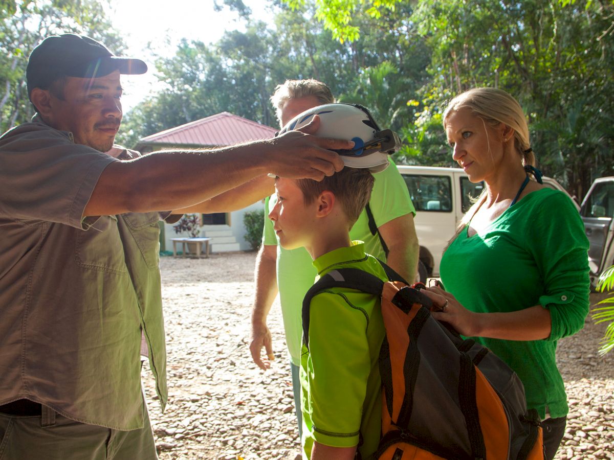 A man is placing a helmet on a boy while two adults, one man and one woman, watch in a forested area with a white van in the background.