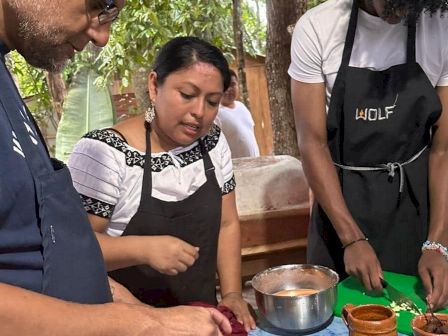 Three people are preparing food at a table outdoors. They are wearing aprons and working with various ingredients and utensils.