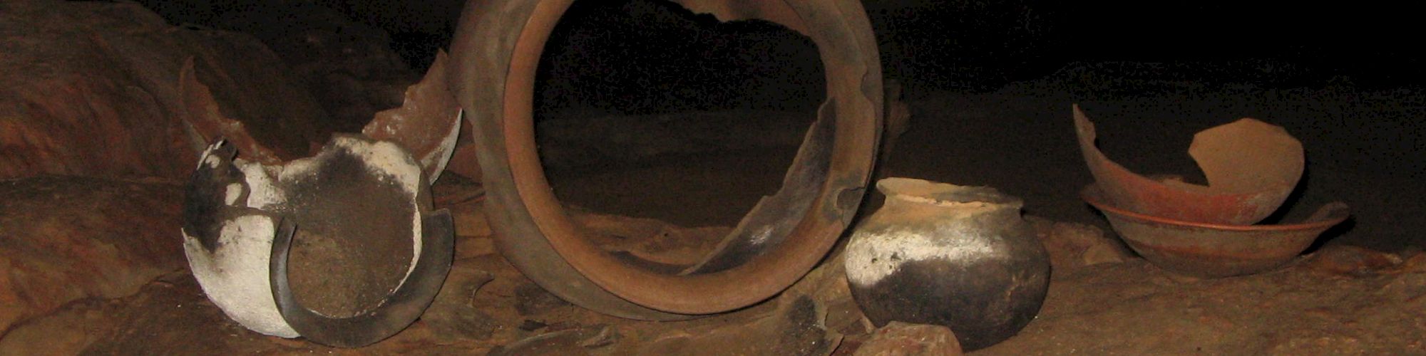 The image shows a cave floor with broken pottery artifacts scattered across the surface, including partial vessels and a large broken ring.