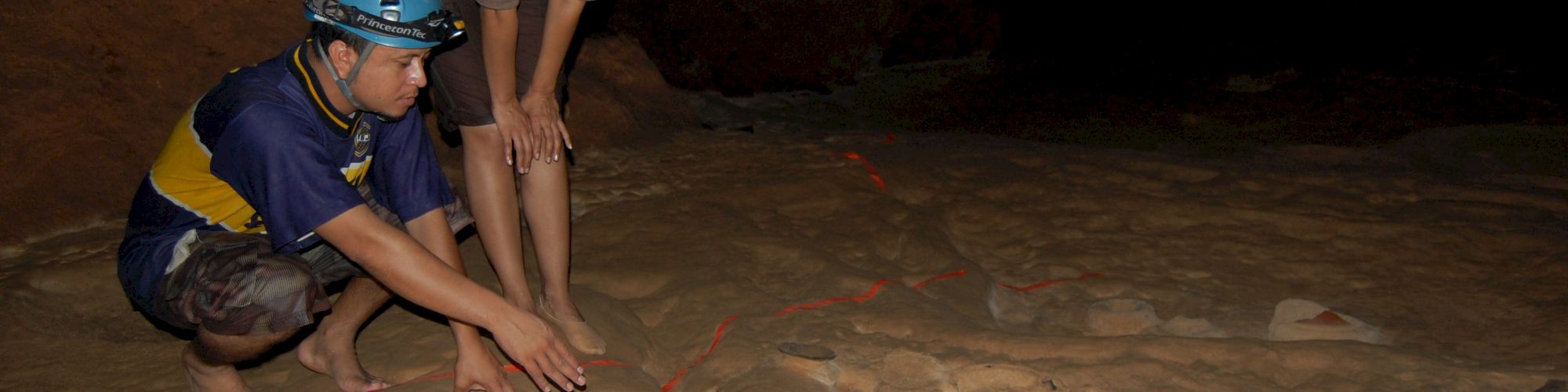 Two people wearing helmets are examining a large rock surface with visible carvings, in a dimly lit environment.