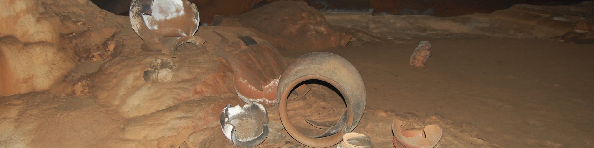 The image shows archeological pottery and artifacts arranged on a cave floor, surrounded by rock formations in a dimly lit environment.