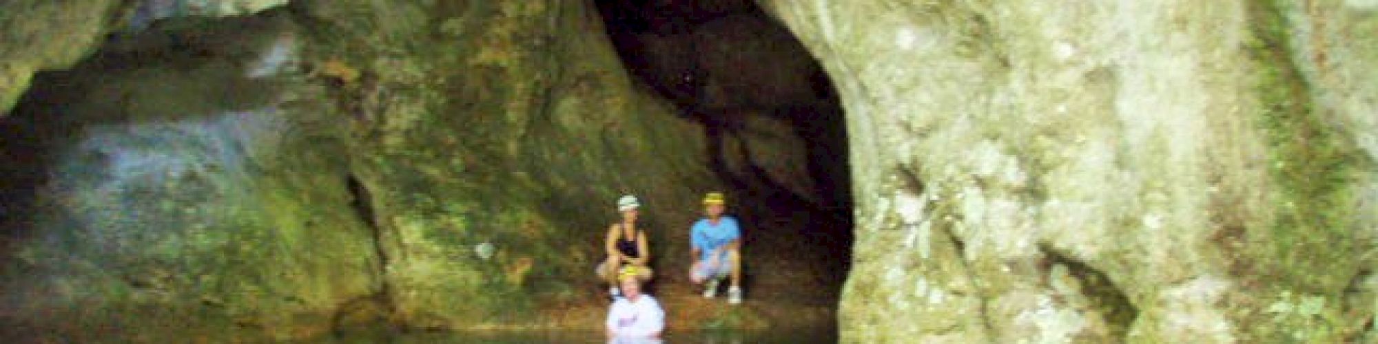 Three people standing in shallow, clear water inside a large cave with rugged rock walls.