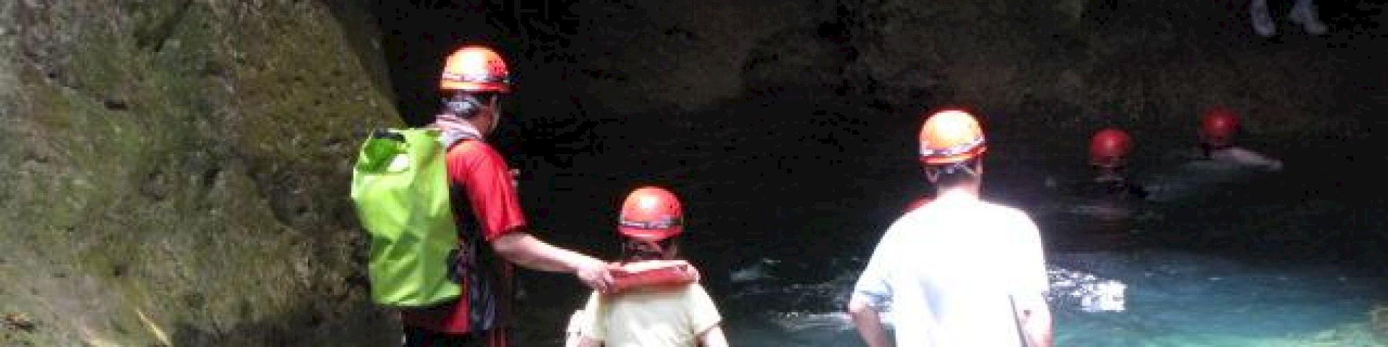 People wearing helmets and safety gear are wading through water in a cave with rocky walls and natural light.