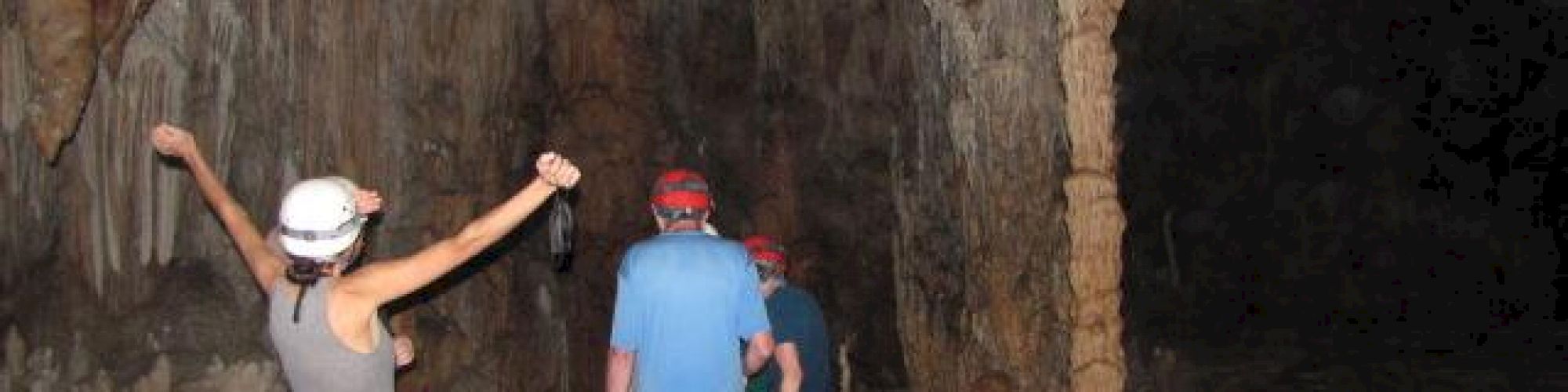 Several people exploring a cave with stalactites and stalagmites, wearing helmets and casual clothing. One person has their arms raised.