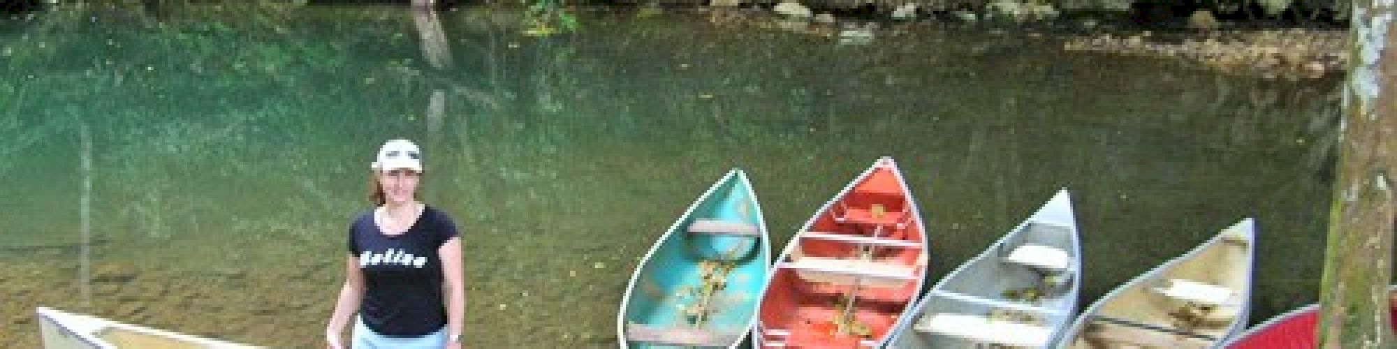 A person stands beside five colorful canoes on a riverbank, surrounded by lush greenery and calm water in the background.