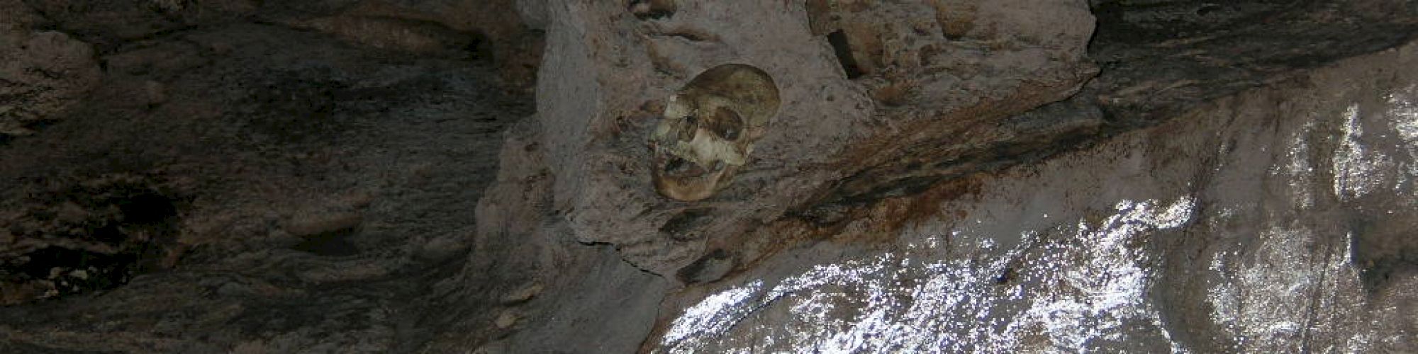 The image shows a cave wall with a human skull embedded in the rock. The scene gives an eerie and ancient feeling, suggesting historical or archaeological significance.