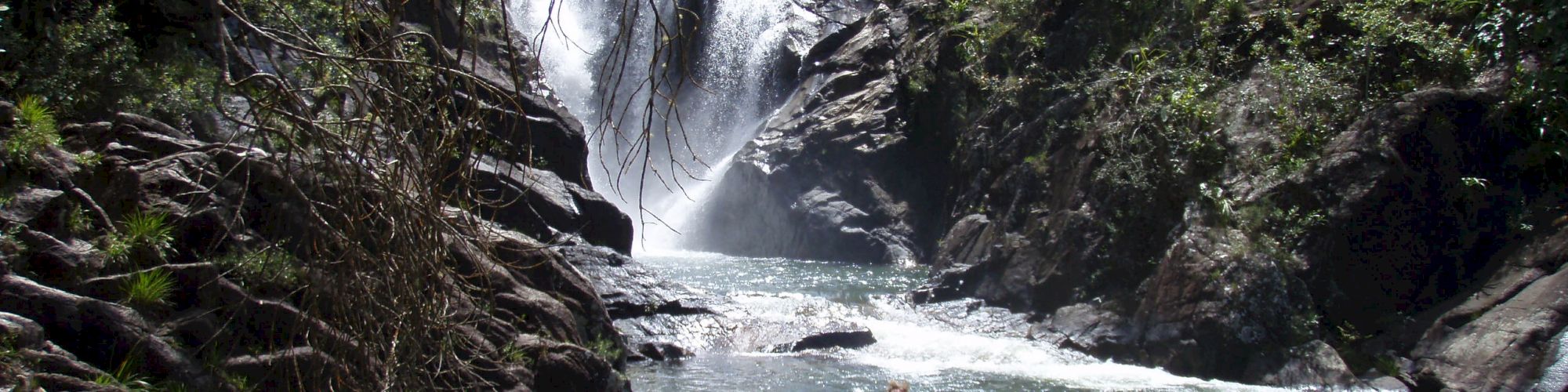 A person is swimming in a river pool near a cascading waterfall surrounded by rocky terrain and lush greenery.