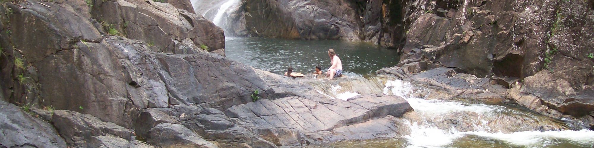 A person sits on rocks near a small waterfall and natural pool surrounded by rugged terrain and greenery.