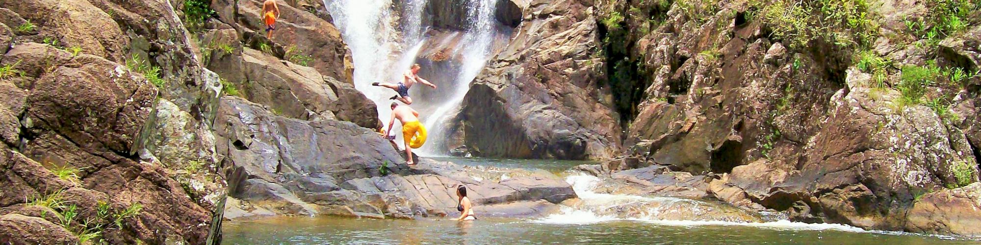 People swimming and jumping into a natural pool at the base of a waterfall surrounded by rocks and lush greenery.