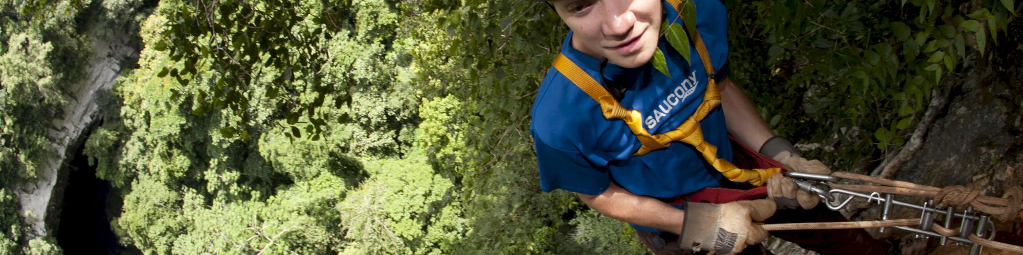 A person in climbing gear ascends a steep, rocky cliff, surrounded by dense greenery in a forested area.