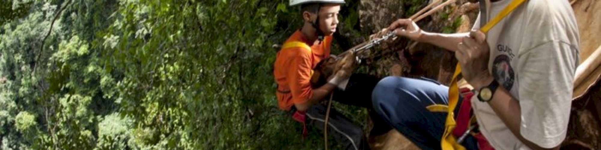 Two people are rock climbing with helmets and harnesses in a forested area. One person is assisting the other on a cliffside.