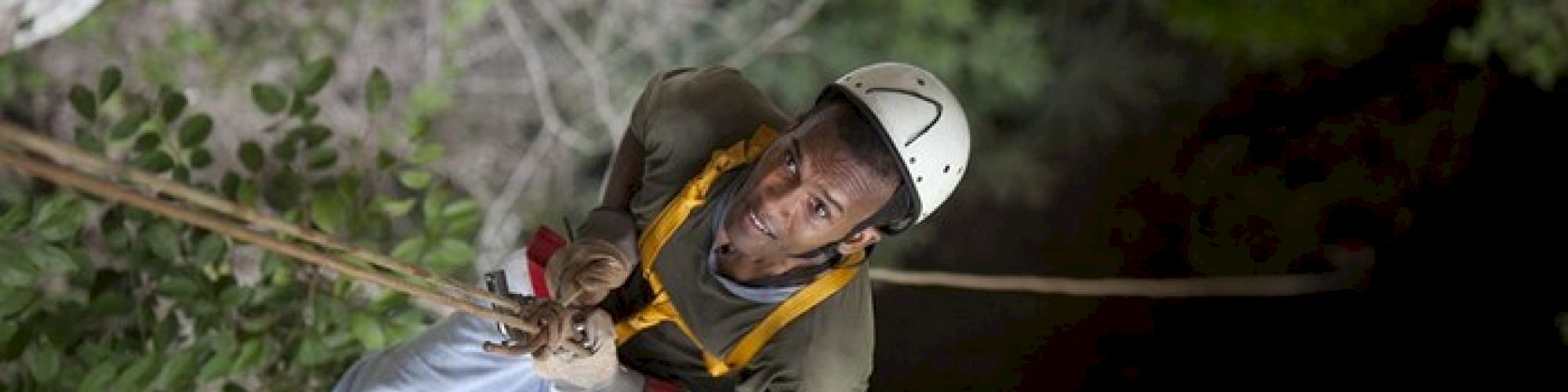 A climber wearing a helmet and harness is ascending a steep rock face, surrounded by greenery.