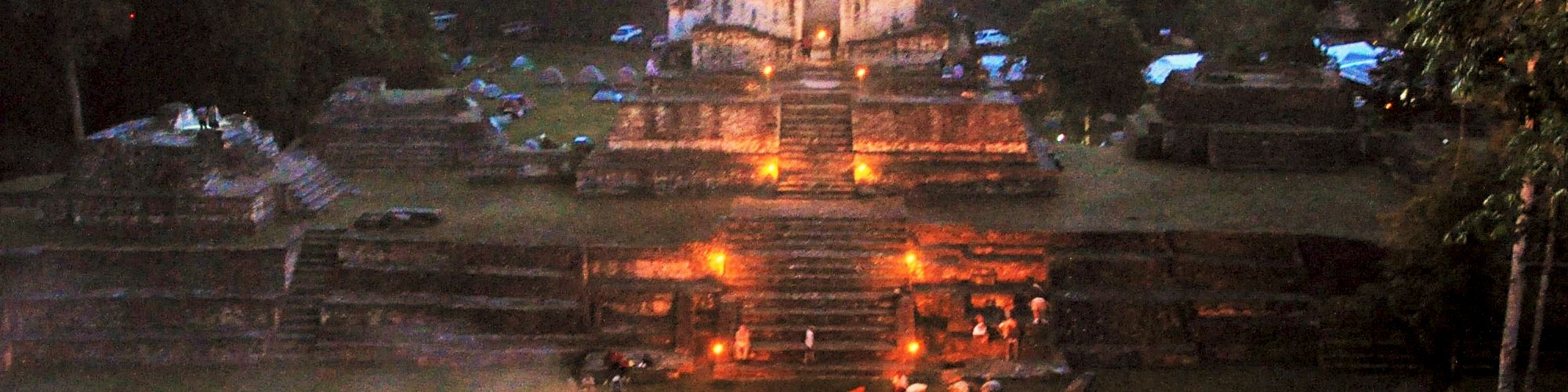 A nighttime scene of an ancient ruins site, with people gathered around a fire in front of illuminated steps against a backdrop of trees.