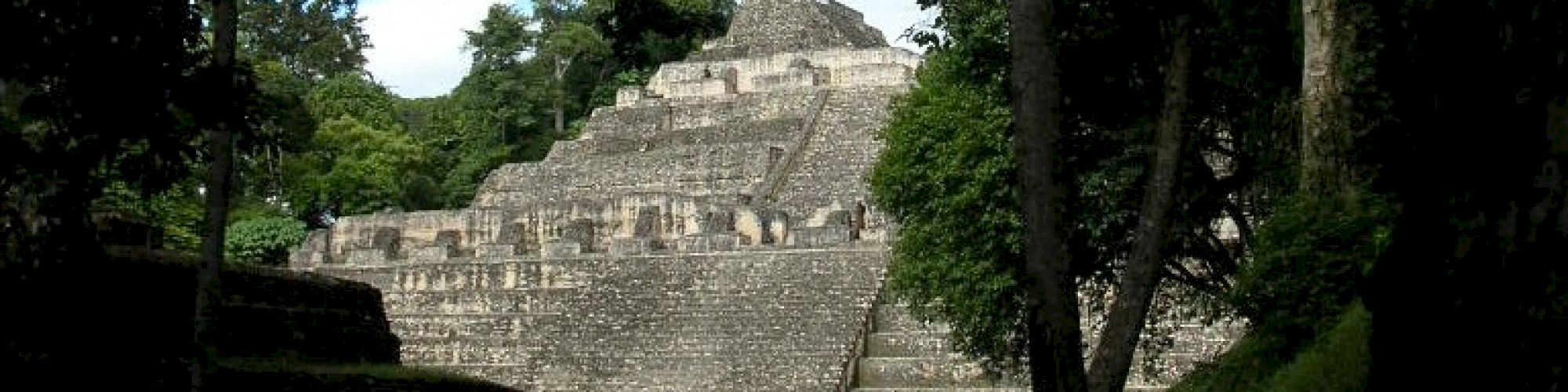 The image shows an ancient stone pyramid structure surrounded by lush trees in a jungle-like setting, possibly a historic ruin site.