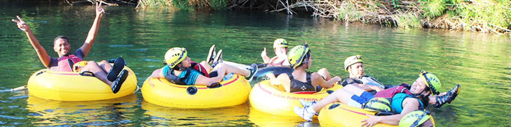 People wearing helmets and life vests are floating on yellow inner tubes in a river, surrounded by lush greenery.