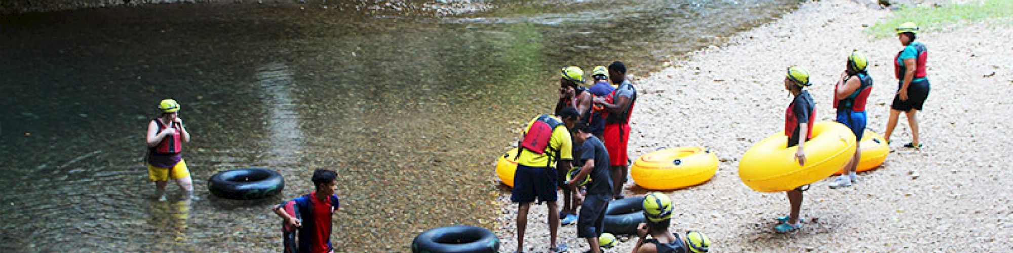 A group of people in life vests and helmets prepare for tubing at a river near a cave entrance, carrying inflatable tubes and standing on a rocky shore.