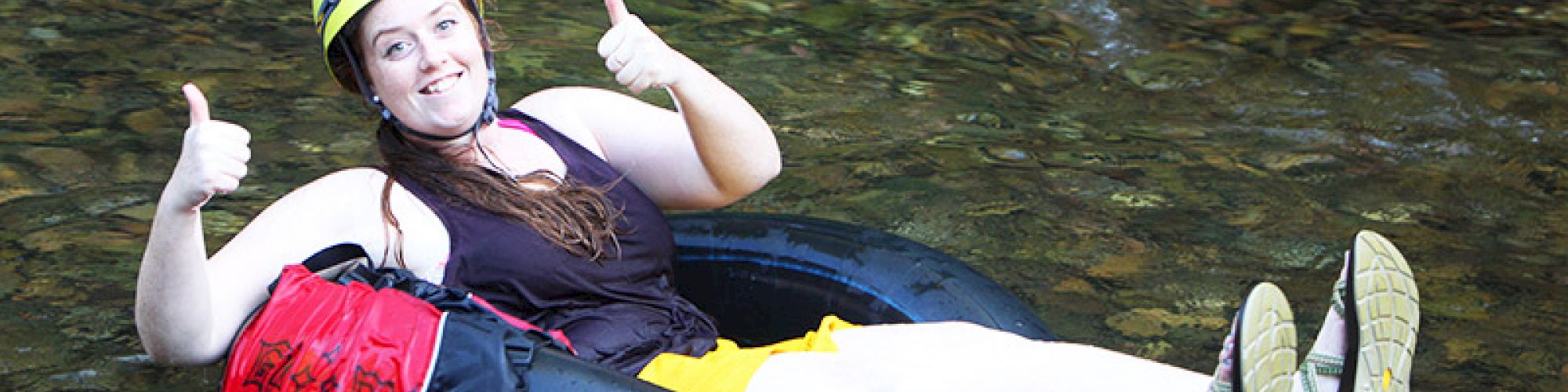 A person in a yellow helmet gives two thumbs up while tubing in a river, wearing a life vest and a black inner tube, with a bag attached.