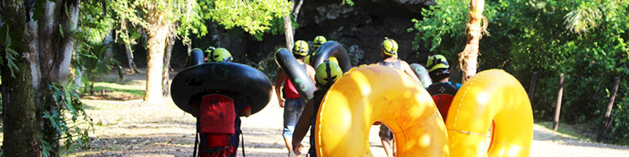 People walking through a forested area carrying inflatable tubes and wearing helmets, preparing for a tubing adventure.