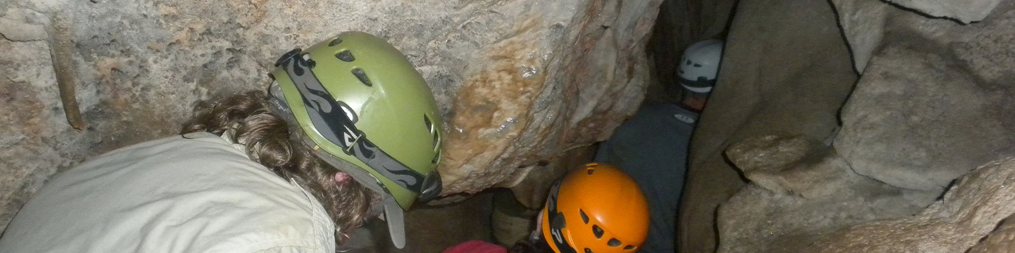 Several people equipped with helmets are navigating through a narrow cave passage.