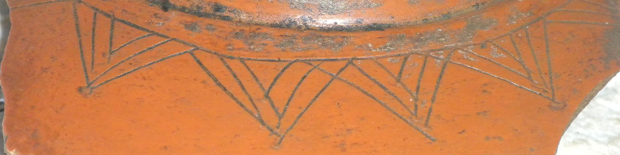 The image shows a piece of pottery with a geometric design etched near its rim, resting on a textured surface.