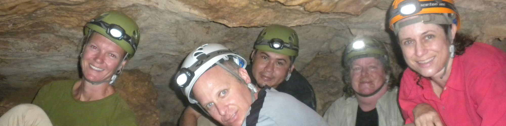 A group of five people wearing helmets and headlamps are in a cave, smiling and posing for the camera.