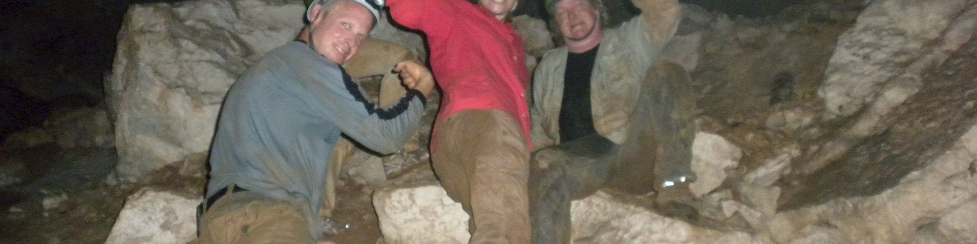 Four people pose flexing their arms in a cave, wearing helmets and head lamps, surrounded by rocks and dirt.