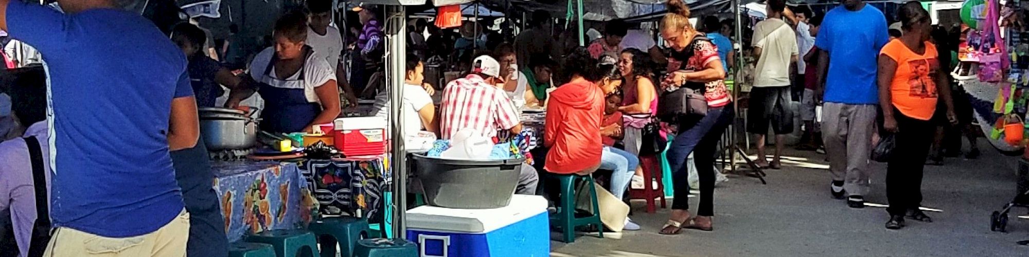 The image shows a bustling street market with people shopping, eating, and interacting under tents and canopies.