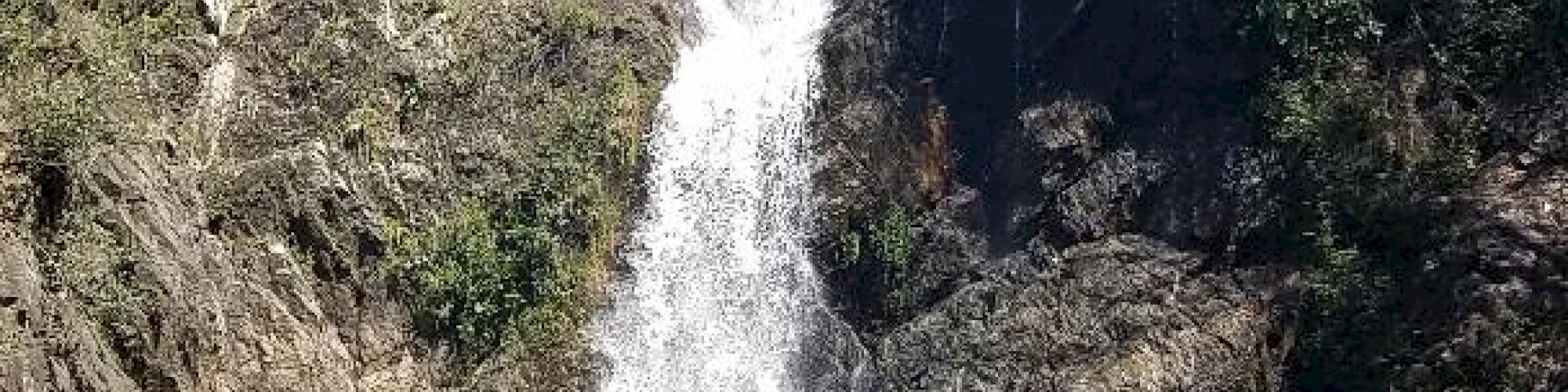 A picturesque waterfall cascades into a serene pool surrounded by lush greenery and rugged rocks on a clear, sunny day.