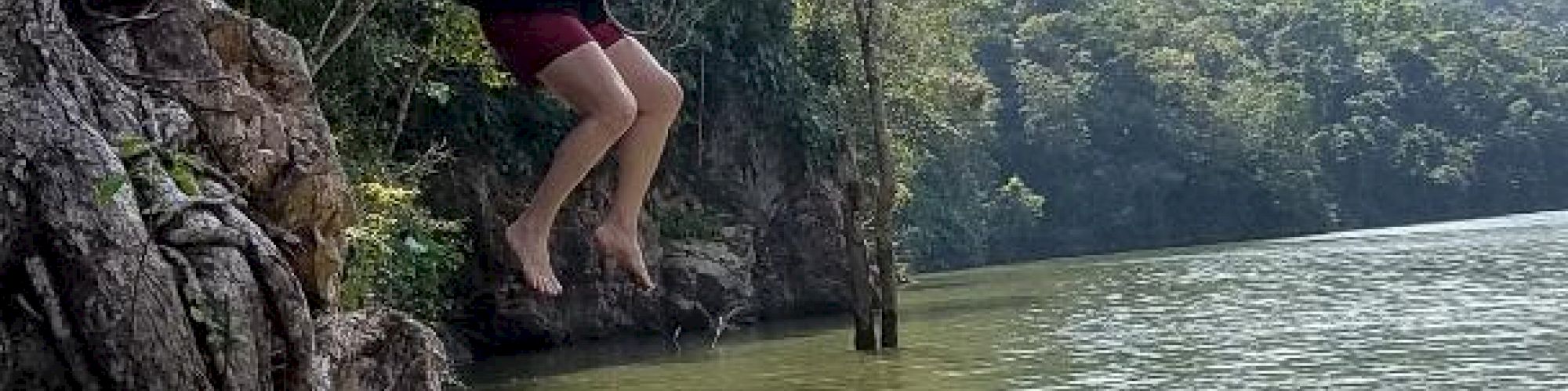 Two people at a rocky lakeside: one person is climbing while the other is mid-jump into water, surrounded by trees and mountainous landscape in the background.