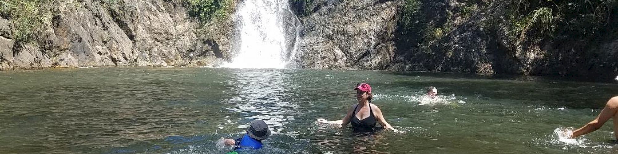 People swimming in a natural pool with a waterfall in the background, surrounded by rocky terrain and greenery.