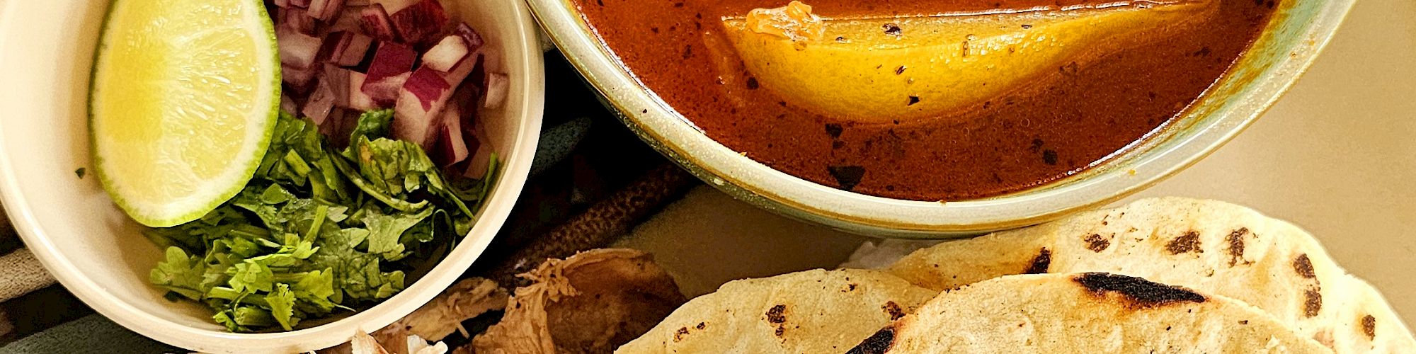 The image shows a bowl of soup, tortillas, shredded meat, and a side of lime, onion, and cilantro on a colorful cloth.