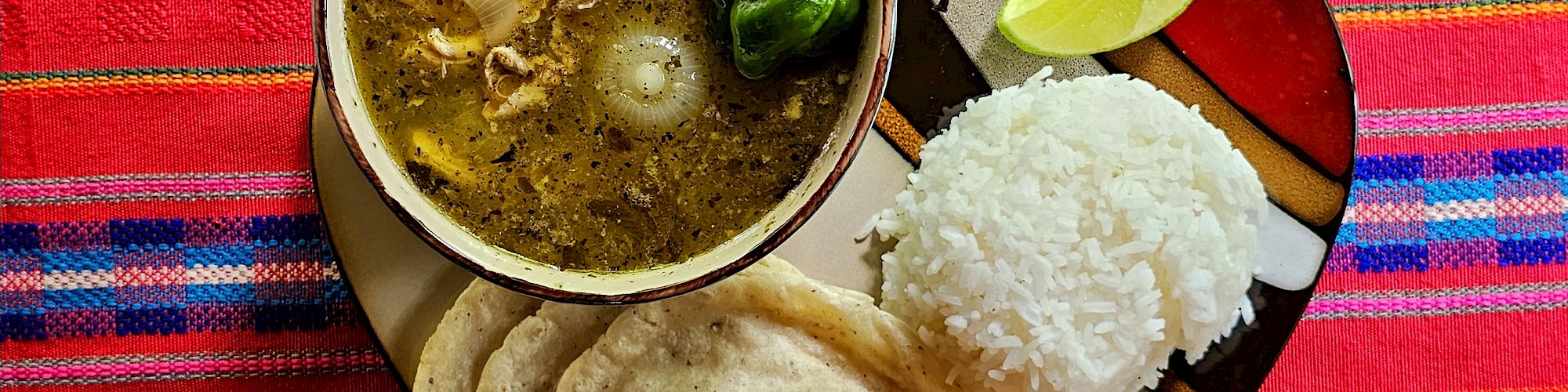 A plate with soup, rice, green chili, lime wedges, and papadums on a colorful cloth.