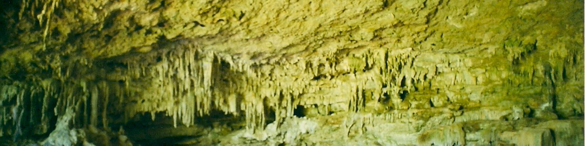 The image shows a large cave with stalactites hanging from the ceiling and a sandy floor below. A person stands inside, highlighting its vastness.