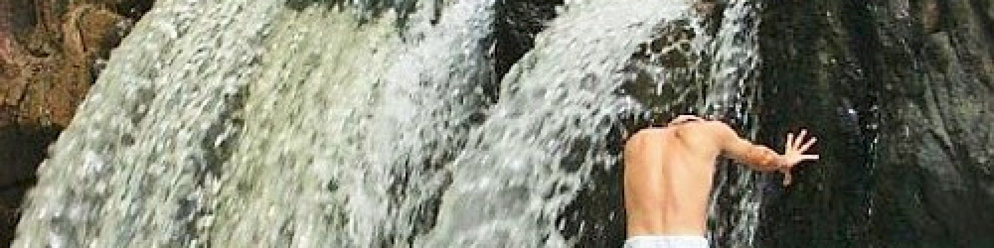 A person in white shorts is standing under a small waterfall with their back to the camera, appearing to be touching the rock wall.