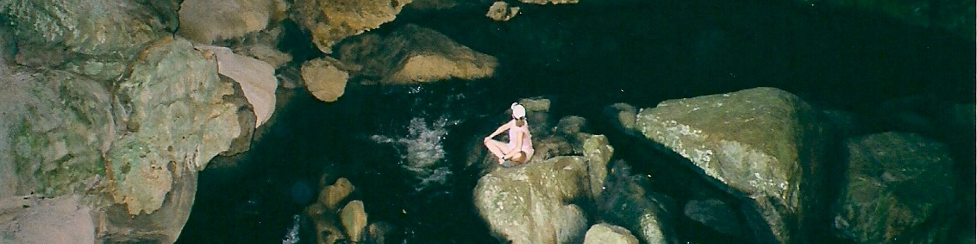 The image shows a person sitting on rocks in a dark, rocky area with water flowing around them, possibly inside a cave.