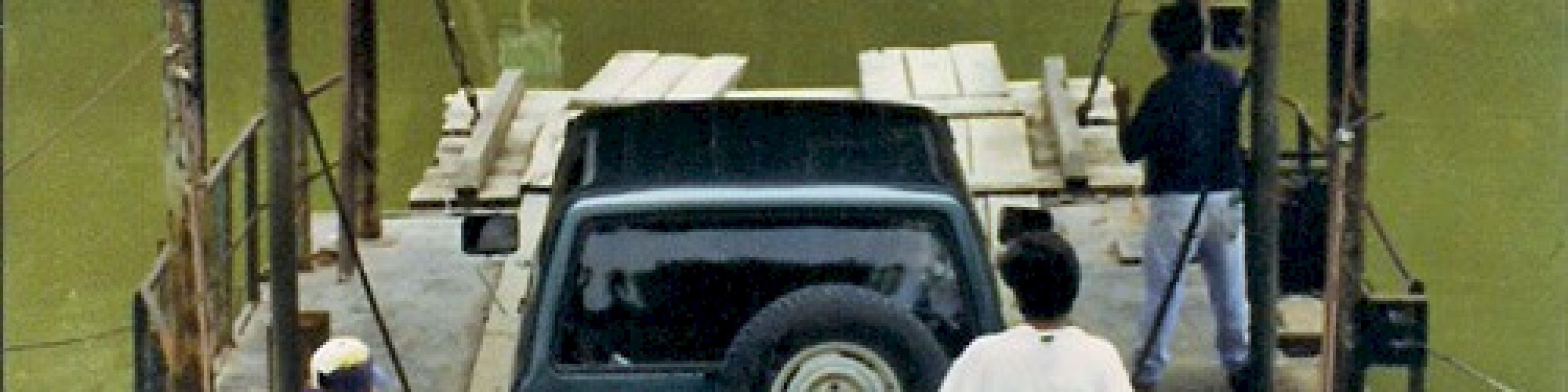 Three people are operating a makeshift ferry transporting a vehicle across a calm body of water.