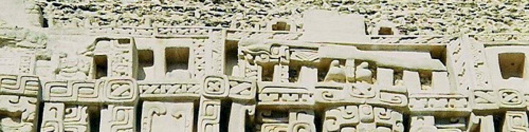 This image shows an ancient stone structure with intricate carvings, possibly Mayan, with two people standing in front of it under a clear blue sky.