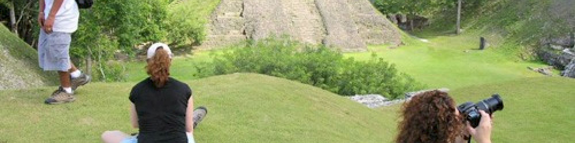 Three people are on grass overlooking ancient ruins; one is taking photographs, another is sitting, and the third stands nearby.